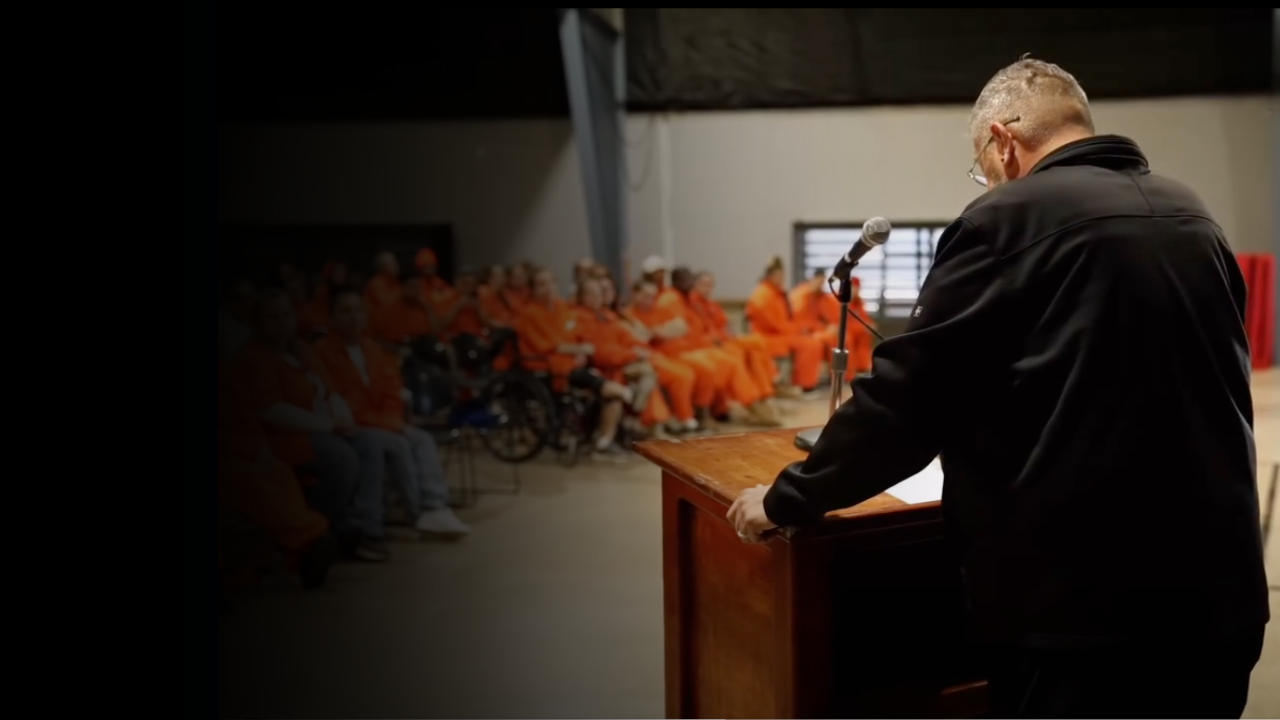 Person at a podium addressing a group of people in orange jumpsuits in a room.